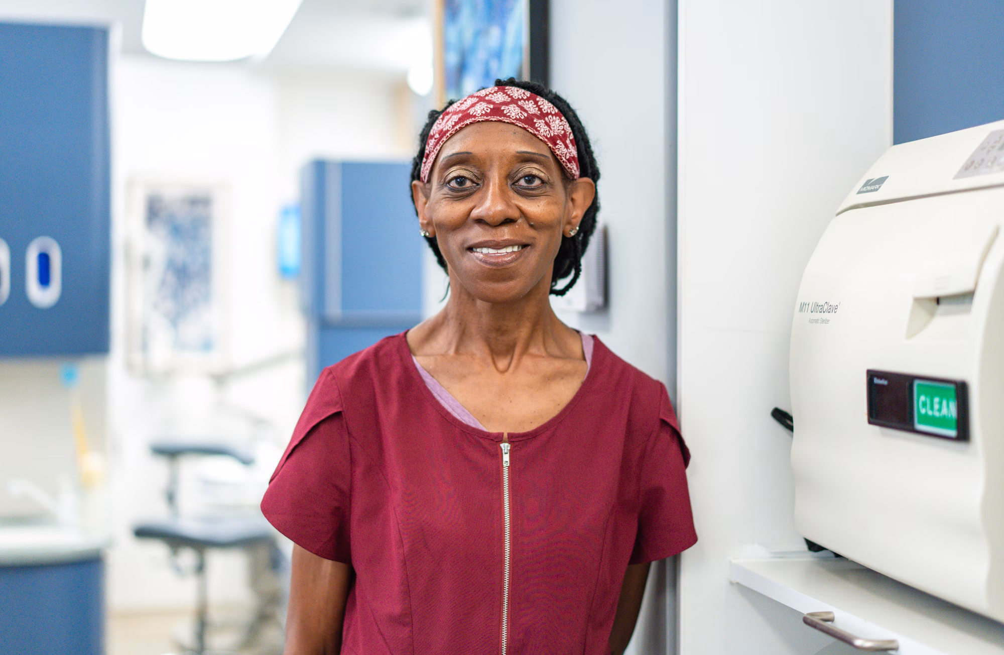 A smiling woman in a maroon scrub top and patterned red headband stands in Whiteman Dental Associates’ Brookline, MA office.